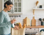 A woman looks at her grocery bill in a kitchen. She is surrounded by bags of groceries.