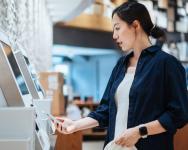 A woman shops at a self-checkout machine. She holds her phone to it to pay with her digital card. She is carrying a shopping bag.