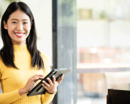 A woman stands smiling. She holds a tablet computer. She is wearing a bright yellow shirt.